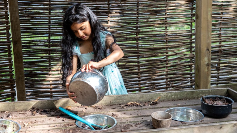 A girl in a blue dress plays with pots and pans, creating something in the mud pie kitchen.